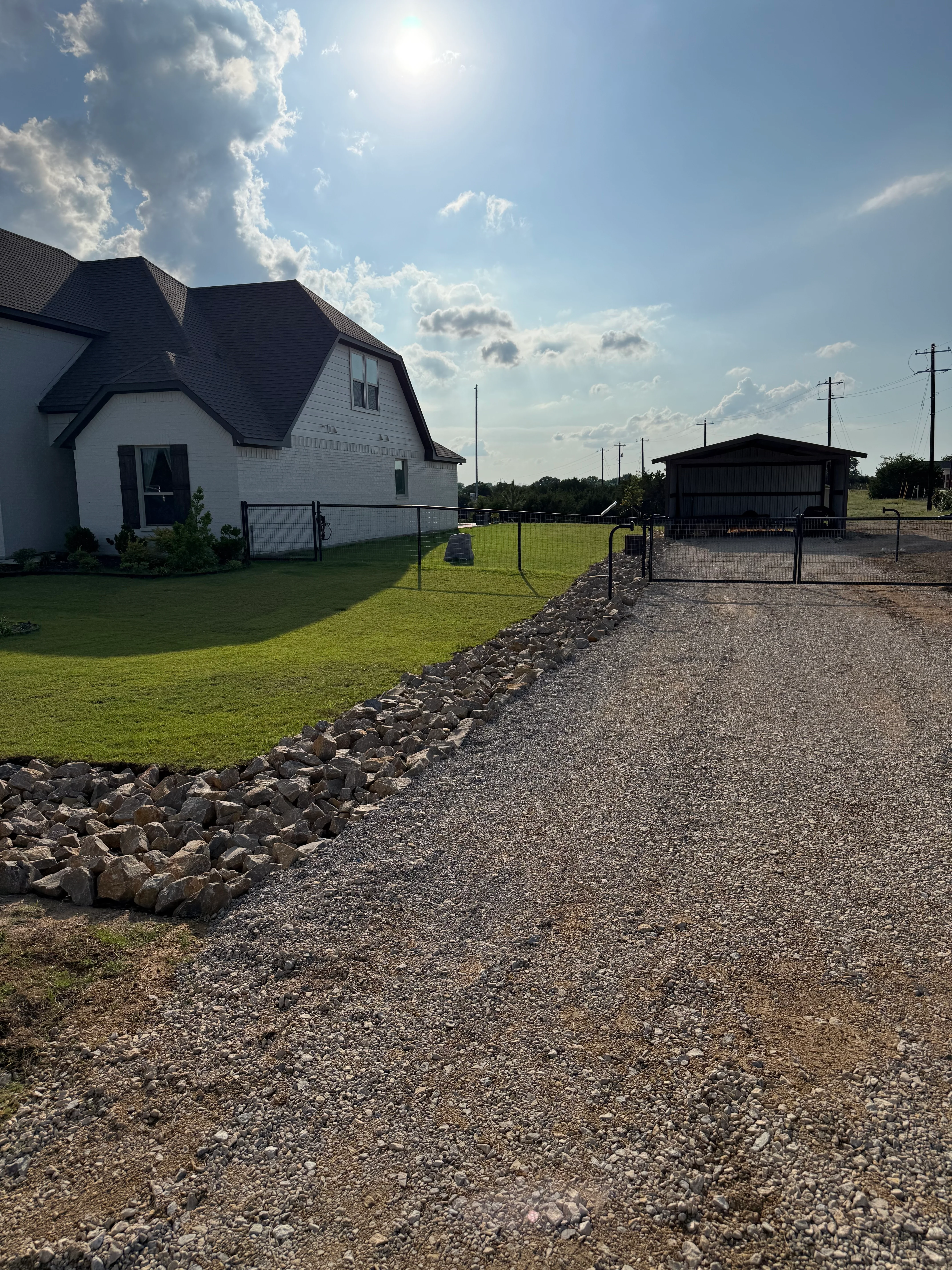 Gravel driveway with rock edging leading to white farmhouse with metal gate fence