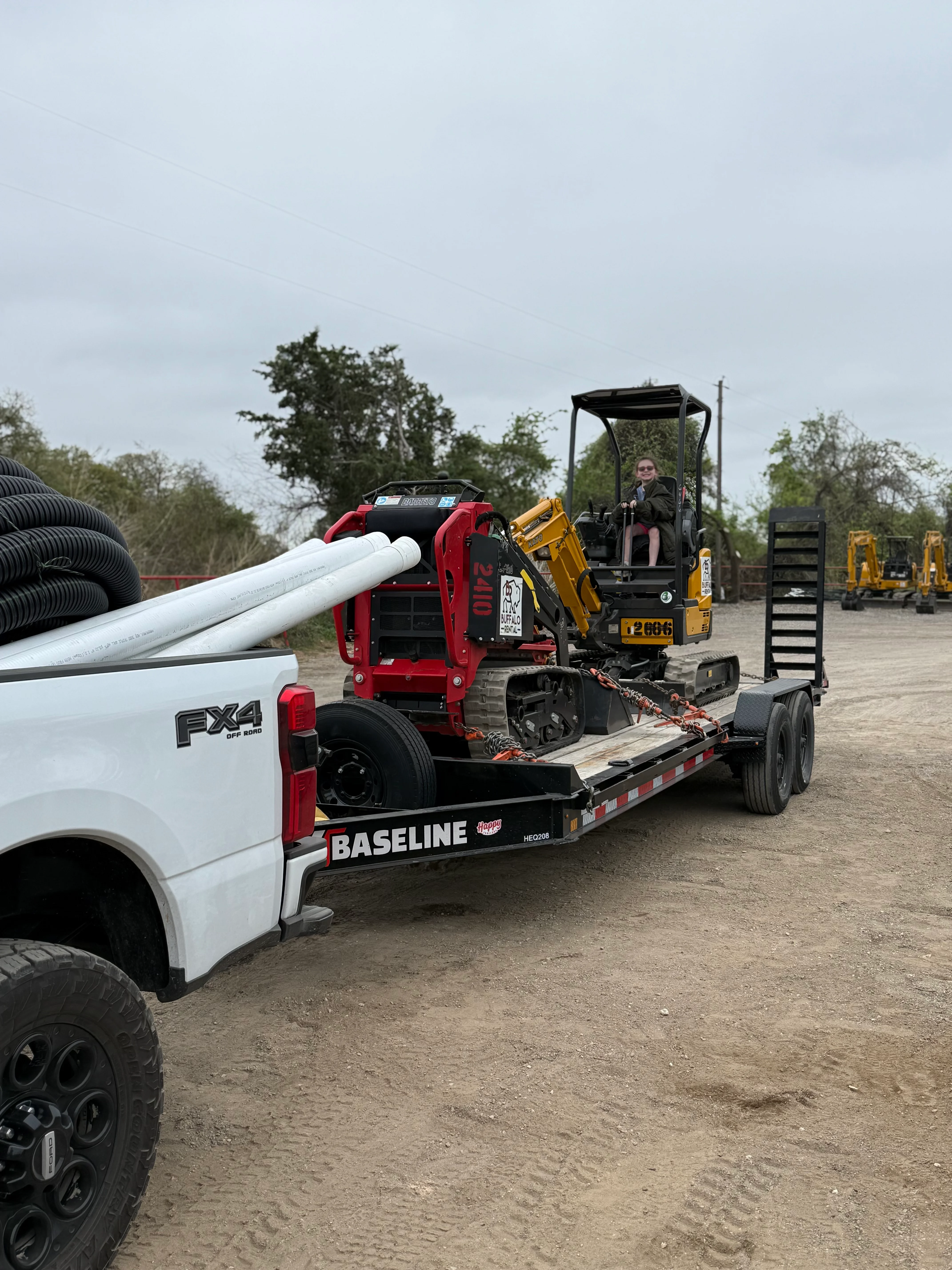 White Ford truck with trailer hauling red mini excavator and PVC pipe at equipment yard