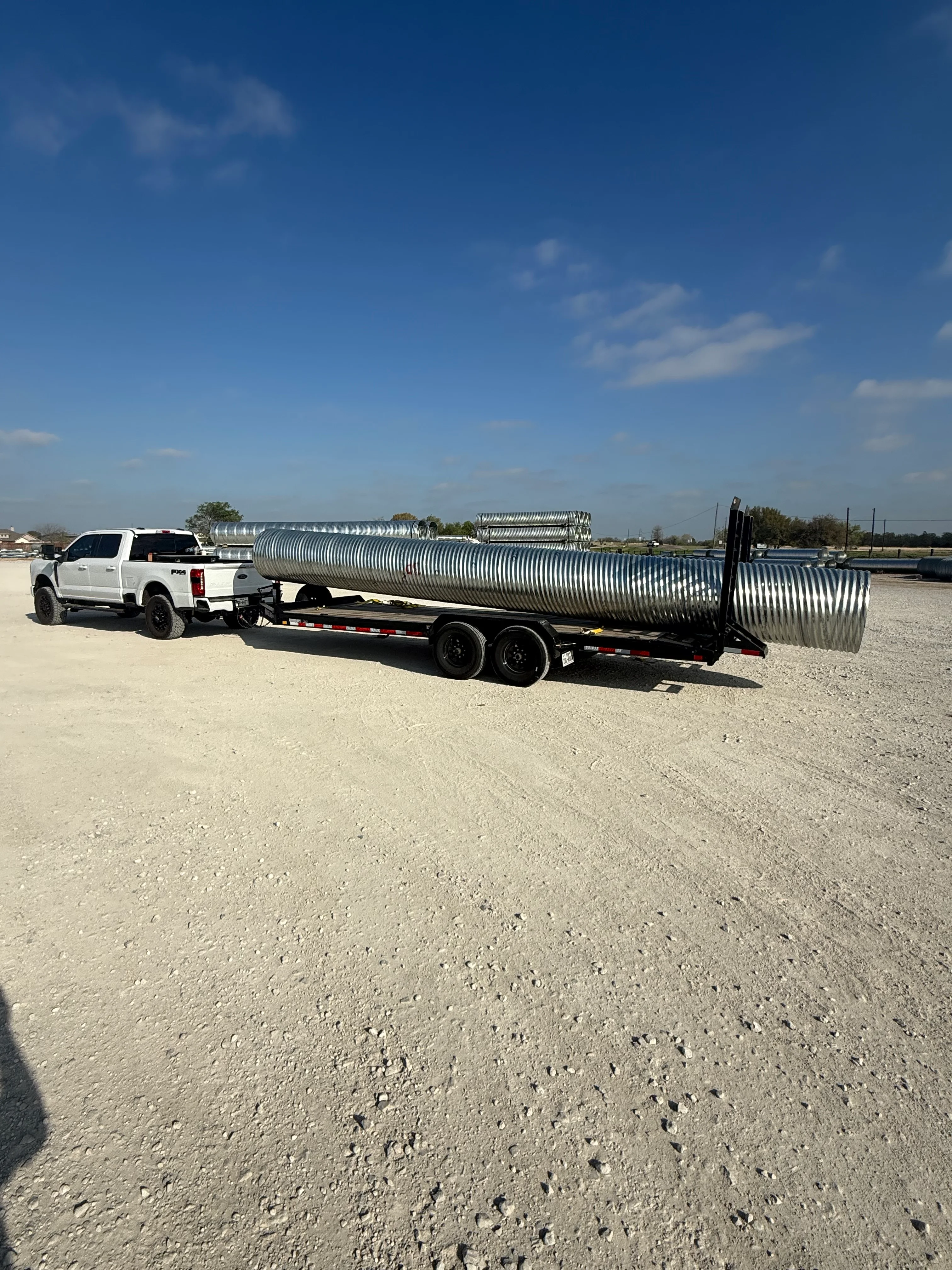 White pickup truck hauling large corrugated metal culvert pipes on flatbed trailer
