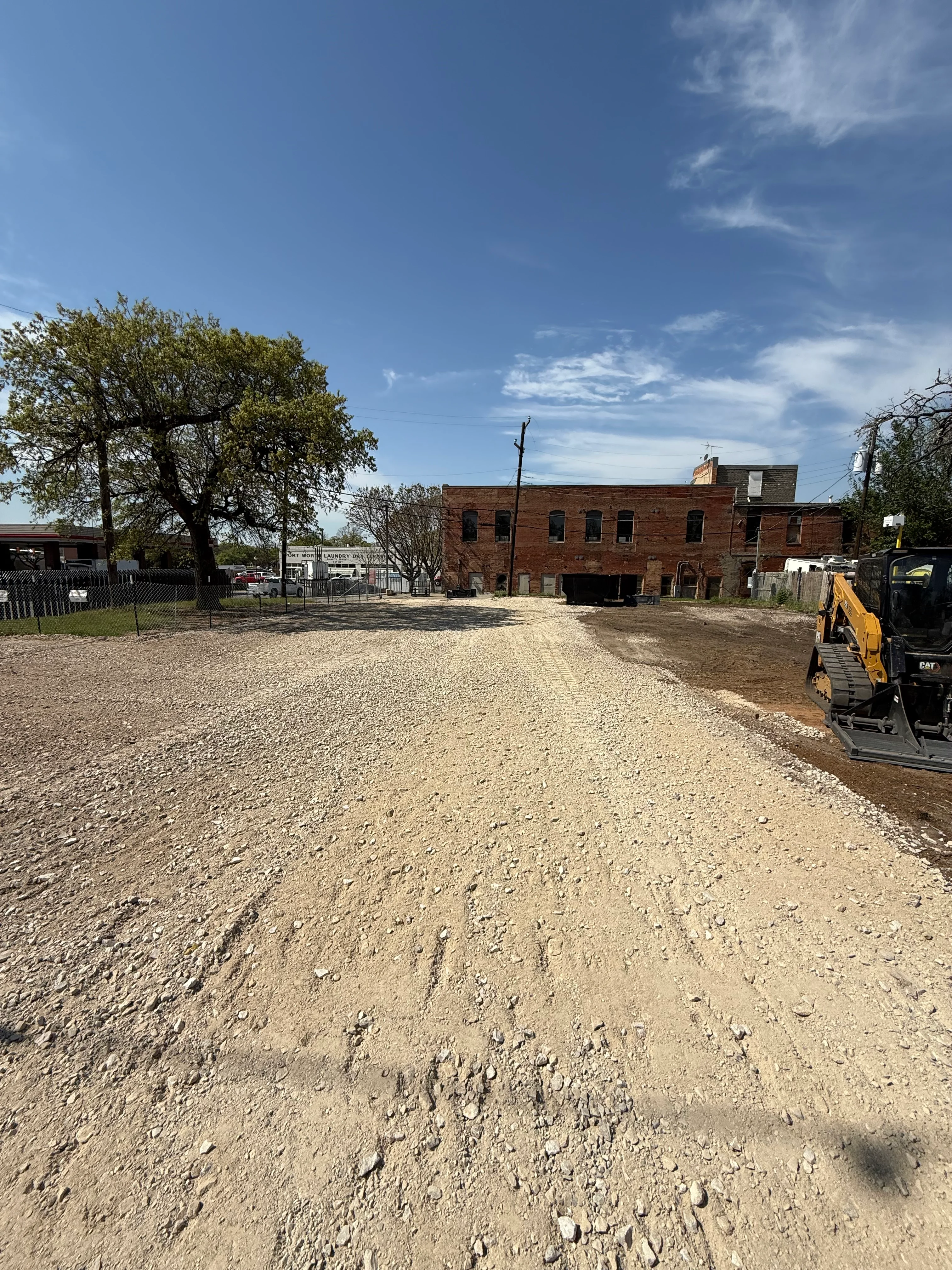 Graded gravel lot with compacted base, brick industrial building, and yellow excavator on site